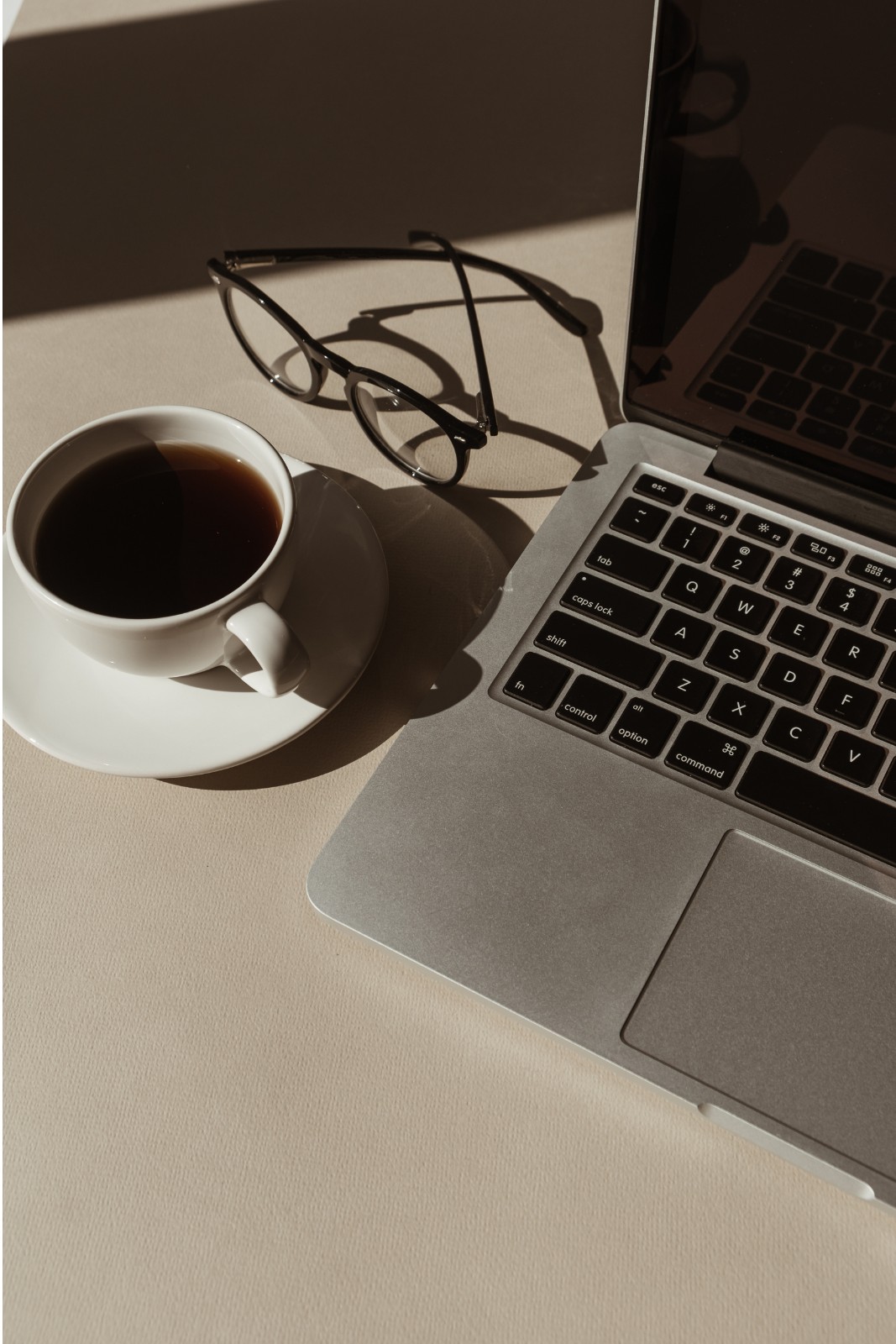A cup of black coffee and reading glasses beside a laptop in morning light.